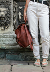 woman with white pants and black belt holding a leather backpack in hand. close up 