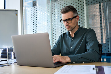 Focused middle aged business man typing on computer working in modern office. Serious busy male manager executive using software laptop analyzing digital tech data doing market research at workplace.