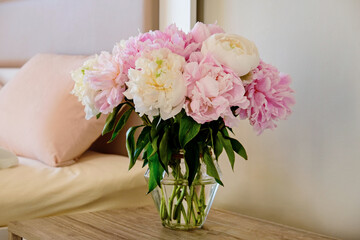Close up shot of the nightstand with a bouquet of peonies in a glass vase near the unmade bed. Good morning concept. Bedroom full of natural light. Copy space for text, background, top view.
