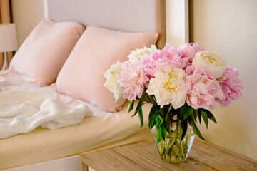 Close up shot of the nightstand with a bouquet of peonies in a glass vase near the unmade bed. Good morning concept. Bedroom full of natural light. Copy space for text, background, top view.