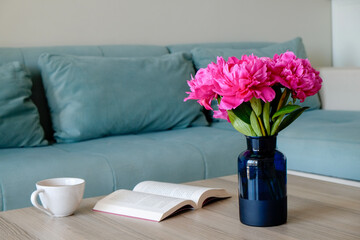 Close up shot of blue glass vase with peony flowers and a notebook with blank pages on wooden table and blue textile couch on background. Simple interior design. Copy space for text.