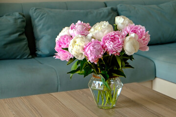 Close up shot of wooden coffee table with bouquet of beautiful white peony flowers in glass vase on foreground and blue textile couch on the background. Copy space for text, close up.