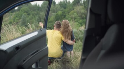 Young father hugging little daughter pointing away talking in countryside meadow in forest. Back view of relaxed Caucasian man enjoying leisure with girl outdoors. Shooting from car