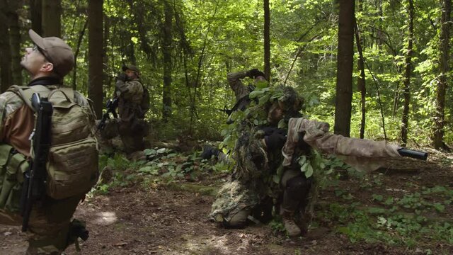 Group of fully equipped soldiers in camouflage armed with rifles, machine gun, shotgun observing approaching of helicopter, giving hand signals to evacuation transport, using radio call