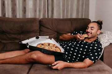 Black man on living room sofa eating pizza and watching streaming.