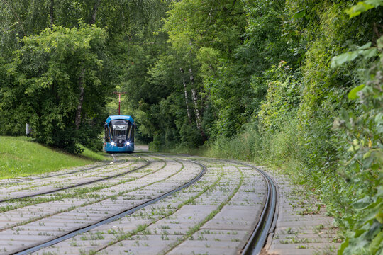 A city or suburban tram on the rails blurs in the distance. Endless curved railway on concrete slabs with sprouted grass. Green arch of trees closes over the tram. Railway disappears into the distance
