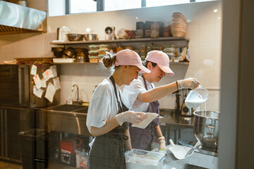 Employees pours milk into bowl making dough with colleague in craft bakery - obrazy, fototapety, plakaty
