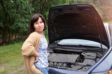 A young woman standing near a broken car with a raised hood is experiencing problems with her vehicle.