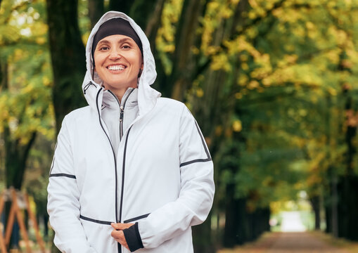 Sincerely smiling Fit athletic woman wearing a modern waterproof running jacket with colorful green yellow autumn park background. Active people concept image.