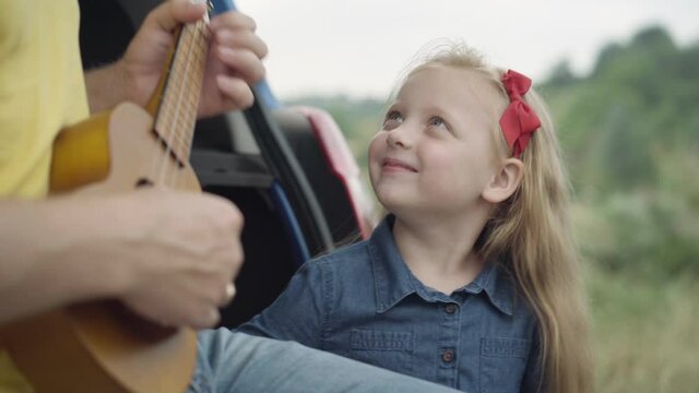 Portrait of smiling charming cute girl admiring unrecognizable man playing ukulele sitting in car trunk. Happy carefree Caucasian daughter enjoying music as father touching strings