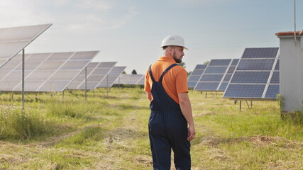 Maintenance assistance technical worker in uniform is checking an operation and efficiency performance of photovoltaic solar panels. Construction engineer walks between solar panels on field station.