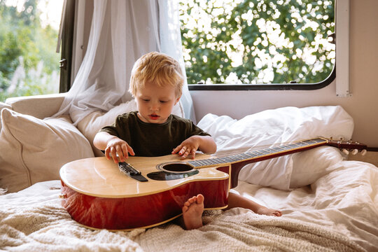 Cute small boy of 2 trying to play guitar sitting alone on bed in trailer rv. Adorable little child entertain hold musical instrument travelling with parents on camper. Childhood and love concept
