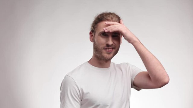 Handsome man touches his face, hair and looks at the camera. Young Caucasian blond curly male straightens his hair in casual clothing posing isolated on white background in studio. Slow-motion.