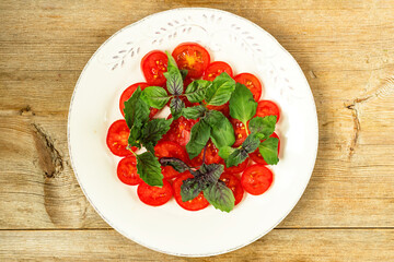 Cut red cherry tomatoes and basil leaves on white plate on wooden table background. Vegetarian vegan food. Healthy food. Clean eating diet. Top view. Flat lay. Close up. Selective focus. Copy space.