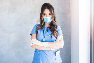 Confident female physician with long hair in blue uniform standing with crossed arms and wearing...