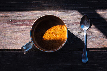 Cup of coffee in a metal mug and a metal teaspoon on a gray-cinnamon wooden surface, light shadow, sunlight of morning coffee, top view