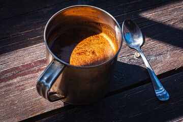 Cup of coffee in a metal mug and a metal teaspoon on a gray-cinnamon wooden surface, light shadow, sunlight of morning coffee, top view