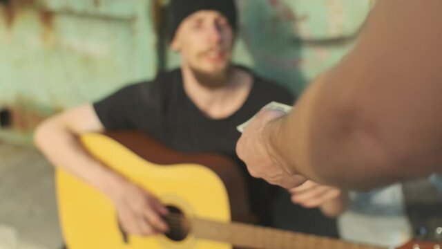 Homeless man on the street by the trash container gets some money playing guitar. broken down person, gotten into poverty, misery, been laid off, fired, sacked. social problems, unemployment concept