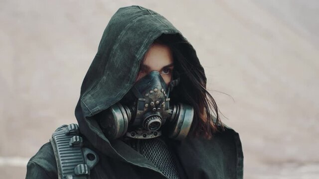 Portrait post apocalyptic woman in grunge clothes hooded mantle and protective gas mask posing by white mountain in desert looking into camera. Future female survivor. Steadicam following shot