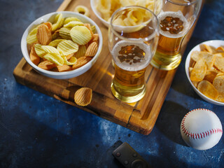 On a wooden tray, two glasses of light beer, onion rings, chips, snacks. There is a remote control and a baseball on the table. Relaxing in the company of sports fans, watching a baseball game on TV.