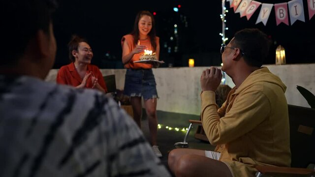 Diversity Asian millennial people friends enjoy outdoor celebration birthday party together with food and drink. Smiling LGBTQ guy excited with birthday cake and blowing birthday candle with happiness
