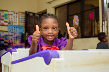 African girl child wearing a purple school uniform, doing thumbs up gestures, while studying and...