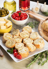 Garlic bread in white square plate on the table with cheese, rosemary, olives and cherry tomatoes.