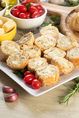 Garlic bread in white square plate on the table with cheese, rosemary, olives and cherry tomatoes.