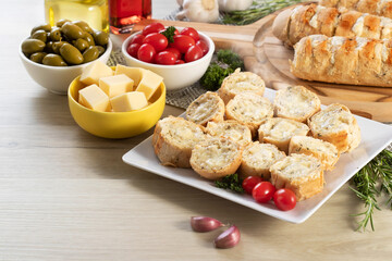 Garlic bread in white square plate on the table with cheese, rosemary, olives and cherry tomatoes.