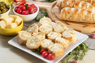 Garlic bread in white square plate on the table with cheese, rosemary, olives and cherry tomatoes.