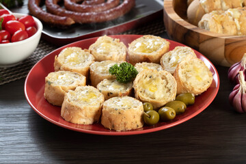 Garlic bread on a red plate on the barbecue table with sausage, cheese, rosemary, olives and cherry tomatoes.
