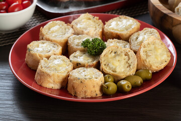 Garlic bread on a red plate on the barbecue table with sausage, cheese, rosemary, olives and cherry tomatoes.