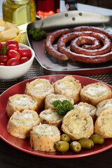 Garlic bread on a red plate on the barbecue table with sausage, cheese, rosemary, olives and cherry tomatoes.