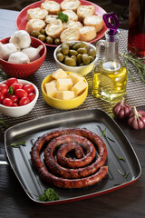 Sausage and garlic bread on a red plate on the barbecue table with appetizers, cheese, rosemary, olives and cherry tomatoes.