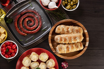Garlic bread on a red plate on the barbecue table with sausage, cheese, rosemary, olives and cherry tomatoes. Top view.