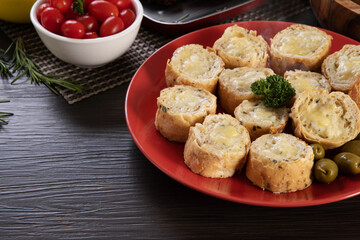 Garlic bread on a red plate on the barbecue table with sausage, cheese, rosemary, olives and cherry tomatoes.