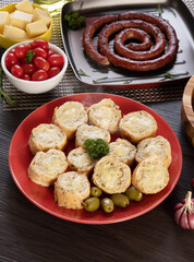 Garlic bread on a red plate on the barbecue table with sausage, cheese, rosemary, olives and cherry tomatoes.