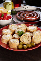 Garlic bread on a red plate on the barbecue table with sausage, cheese, rosemary, olives and cherry tomatoes.
