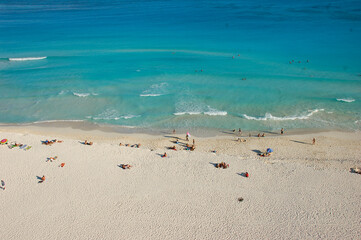 People enjoying a sandy beach on a sunny day with clear blue water and foam-capped waves.