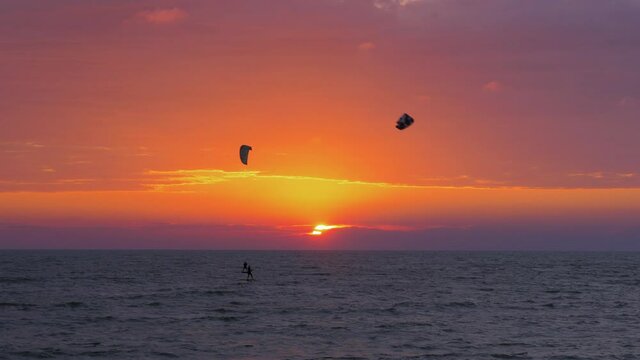 Silhouette of people engaged in kitesurfing in vibrant summer evening just before sun goes down, high contrast beautiful red sunset, Baltic Sea coastline, Karosta beach in Liepaja, wide distant shot