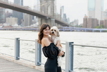 a woman holding her small mixed breed dog at the boardwalk by the hudson river with brooklyn bridge...