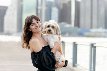 a woman holding her small mixed breed dog at the boardwalk by the hudson river with brooklyn bridge...