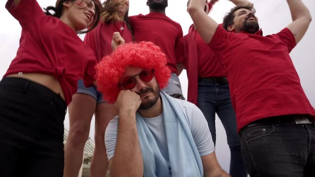 A Group Of Soccer Team Fans Celebrate Victory In The Stands Wearing Red Jerseys While A Fan Of The Opposing Team Mourns In Sadness.