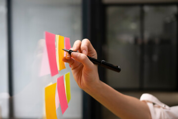 Close up view businesswoman writing creative ideas on sticky notes on glass wall.