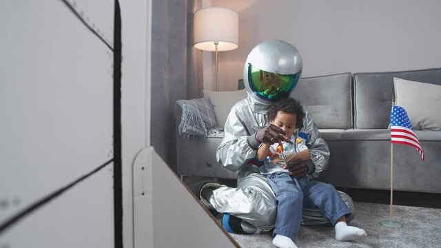 Afro american father and son play in the living room at home, man in a suit of an american astronaut sitting on the floor with her son, boy playing with a toy model of the solar system, the flag of