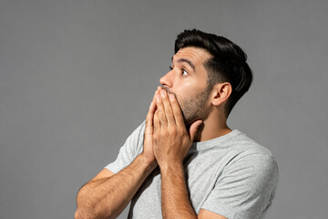 Shocked scared young Caucasian man with eyes popping and hands covering mouth on light gray studio background