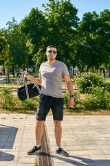 A handsome young Caucasian man with dark hair in sunglasses, a gray T-shirt and black shorts with a sports bag stands against a background of trees. Full-length portrait