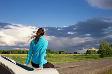 a young girl resting on the trunk of a car against the backdrop of an incipient thunderstorm, during a long journey on a suburban road