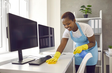 Cheerful smiling cleaner carefully wipes the dust from the table in a bright office. African...