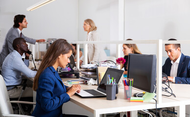 Focused serious young female working with laptop in busy modern open plan office